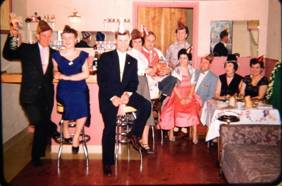 A group of men and women in festive attire and party hats pose together in a vintage-style room, some seated at a bar and others at a decorated table, smiling and celebrating.