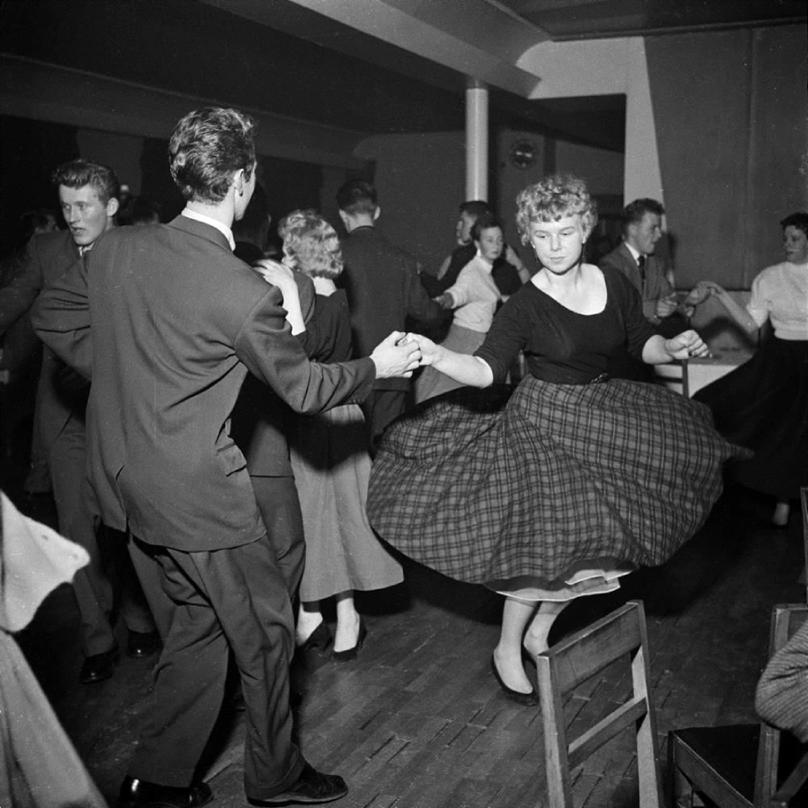 Black-and-white photo of young men and women dancing in pairs, wearing formal 1950s-style clothing. A woman’s plaid skirt flares as she spins, and others dance in the background on a wooden floor.