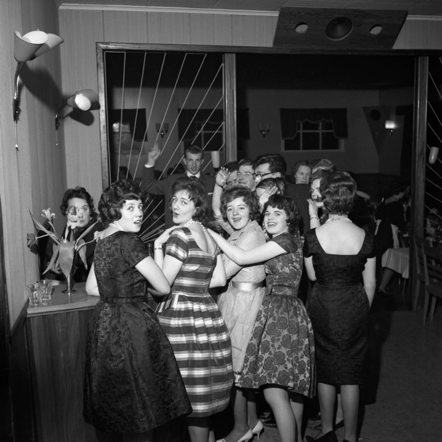 A group of women in vintage dresses smile and pose together at a lively indoor party, while several men in suits wave from the background. The setting appears festive and mid-20th century.