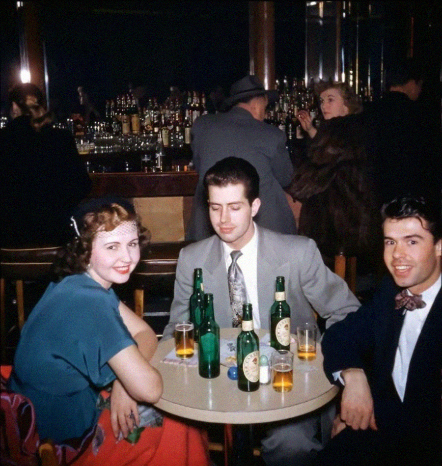 Three people sit at a small round table in a dimly lit bar, with beer bottles and glasses in front of them. Two men in suits and a woman in a blue dress smile, while patrons socialize at the bar in the background.