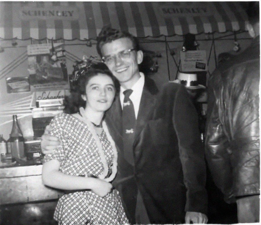 A black-and-white photo of a young woman and man smiling with arms around each other in a lively indoor setting, with a striped awning and shelves with bottles and boxes in the background.