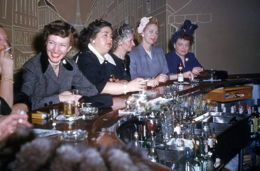 Five women dressed in vintage clothing sit at a bar, smiling and conversing. There are drinks and glasses on the bar, with liquor bottles visible in the foreground. The atmosphere appears lively and social.