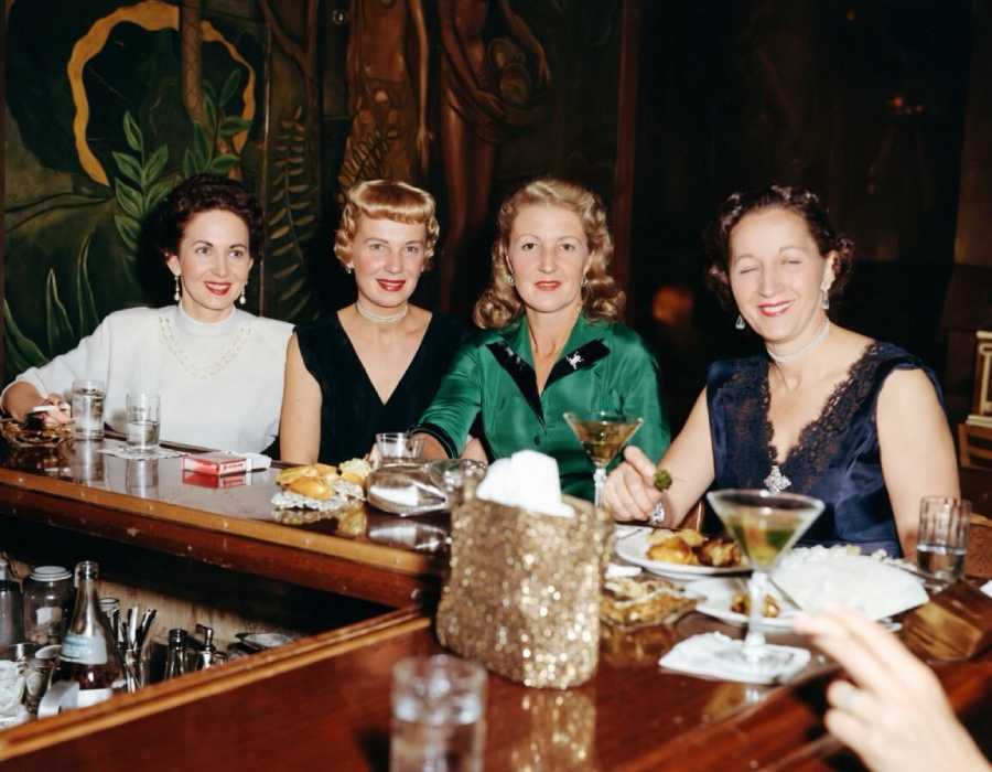 Four women dressed in elegant 1950s-style attire sit at a bar with cocktails, food, and cigarettes, smiling for the camera. The background shows artistic murals and a dimly lit, stylish setting.