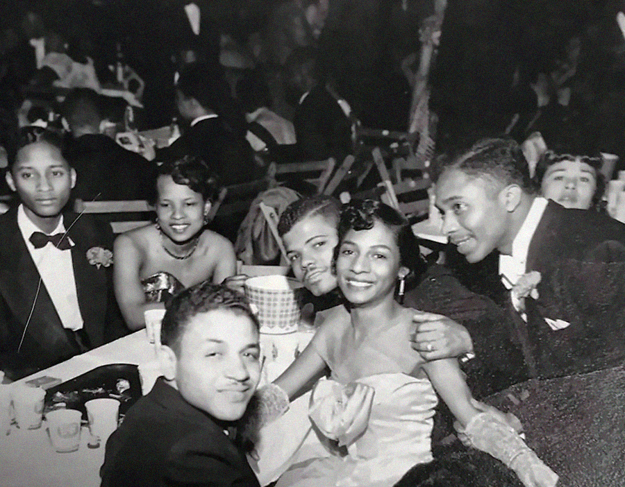 A group of young people dressed in formal evening attire sit around a table at a lively event, smiling and posing for the camera. The setting appears to be a dance or banquet, with others in the background.