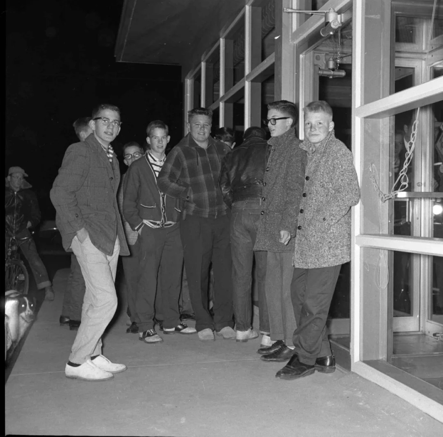 A group of young men in 1950s-style clothing stand outside a glass-fronted building at night, some facing the camera and others turned away, appearing relaxed and social.