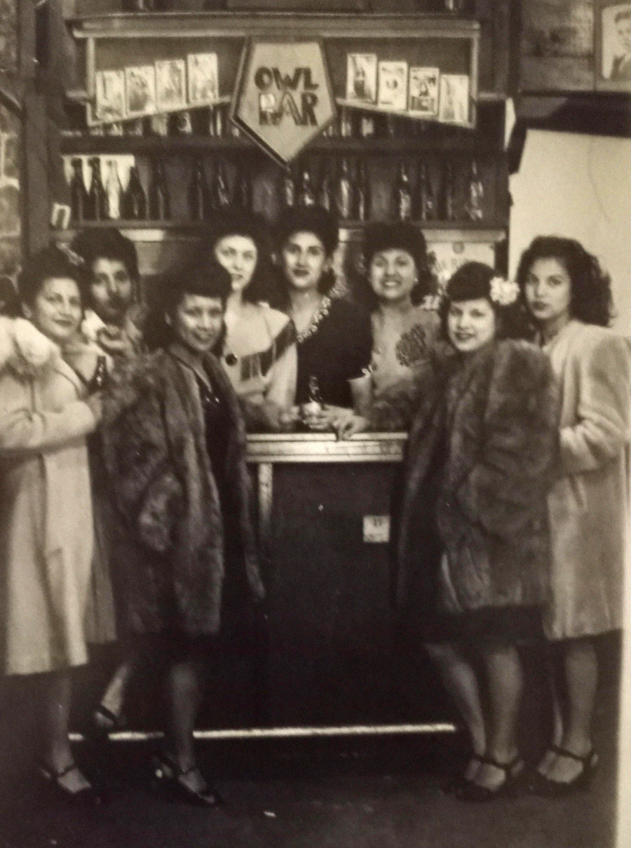 Vintage black-and-white photo of eight women in coats and dresses standing together at a bar with an “OWL BAR” sign above shelves of bottles. They are smiling, posing closely in a cozy, lively setting.