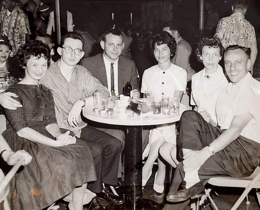 A black-and-white photo shows six adults, three men and three women, sitting around a round table with drinks, smiling at the camera in a busy indoor setting, possibly a bar or club, in vintage attire.