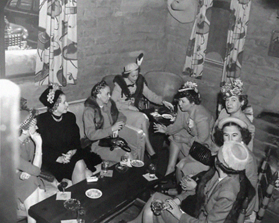 A black-and-white photo shows a group of women in hats and dresses sitting around a table in a cozy room, engaged in lively conversation. Cups, saucers, and cigarettes rest on the table, evoking a social gathering from the past.
