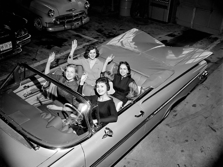 Four women seated in a classic convertible car indoors, smiling and waving at the camera. The car is parked in a garage with other vintage vehicles visible in the background.