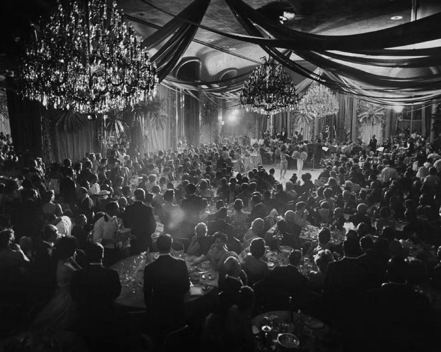 A large crowd in formal attire sits at tables and watches dancers perform in an elegant ballroom decorated with chandeliers and draped fabric. The scene is lively and illuminated by soft, dramatic lighting.