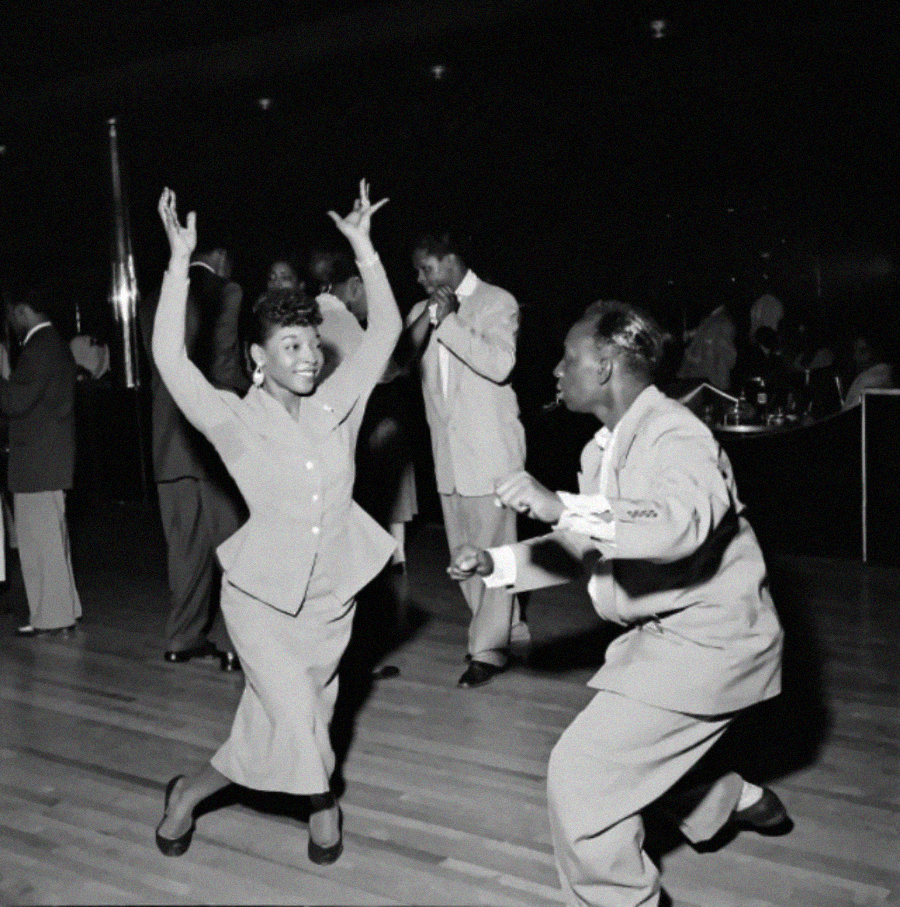 A woman and a man energetically dance together on a wooden floor in a lively, crowded ballroom, while a band performs in the background. The woman raises her arms, both are dressed in suits, and people watch from the sidelines.