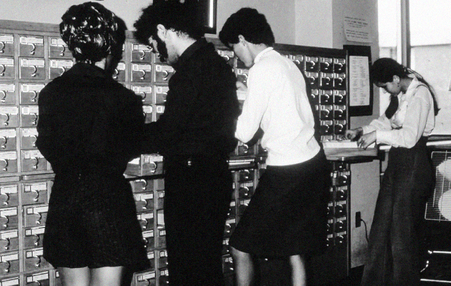 Four people stand in front of large card catalog drawers in a library, searching for information. The photo is black and white, suggesting it was taken decades ago. One person writes at a nearby table.
