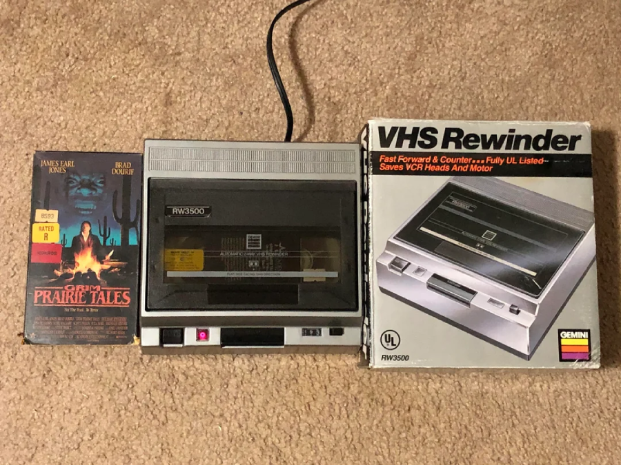 A VHS tape of "Prairie Tales," a VHS rewinder device with a tape inside, and the rewinder’s original box are displayed on a beige carpet.