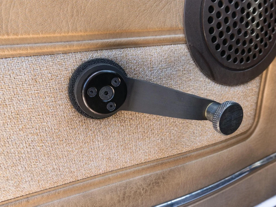 A close-up of a manual car window crank handle on a tan-colored car door panel, with a circular speaker visible in the upper right corner.