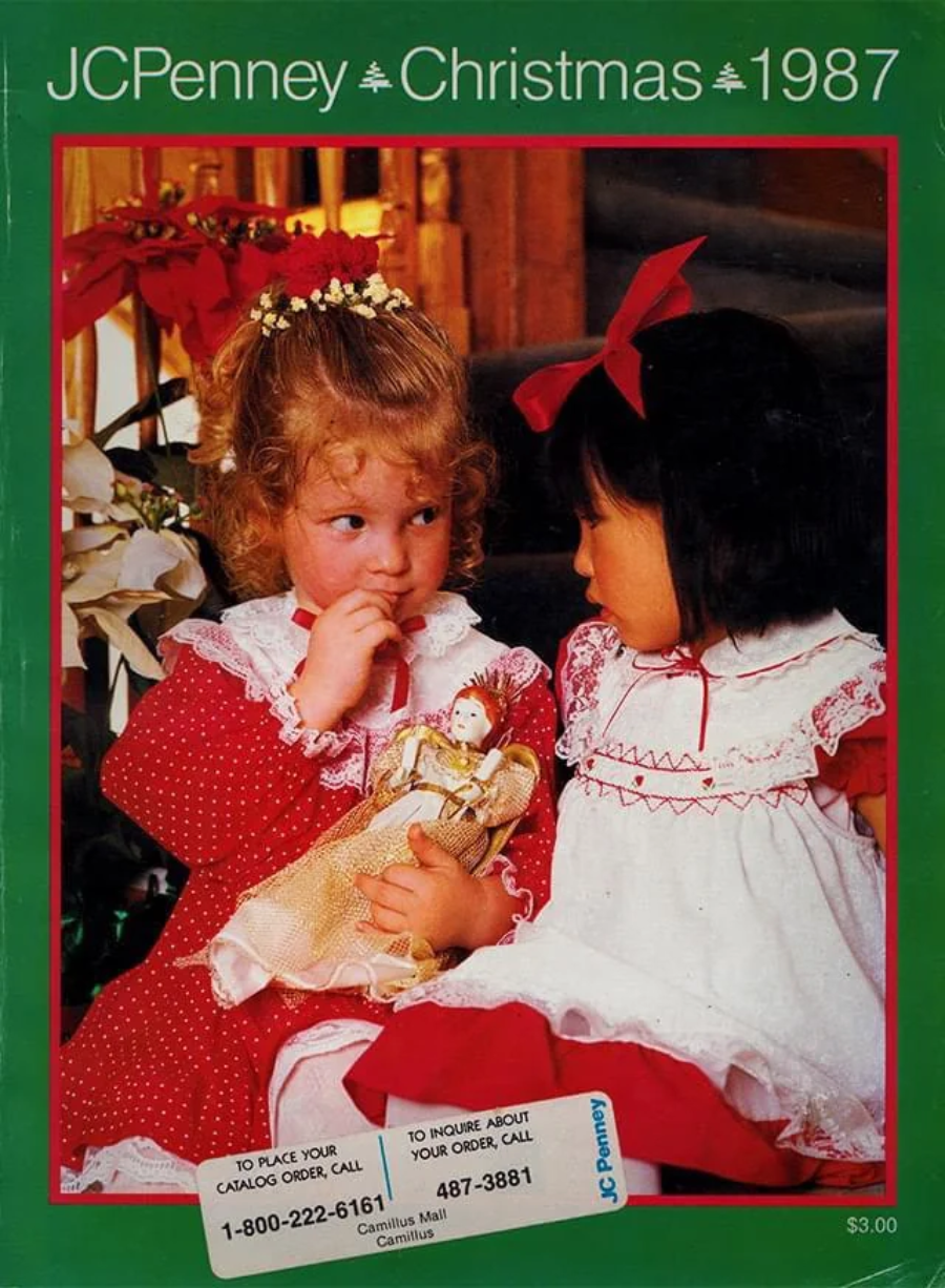 Two young girls in festive red and white dresses sit closely together, one holding a doll. The image is on the cover of a JCPenney Christmas 1987 catalog with green borders and contact numbers at the bottom.