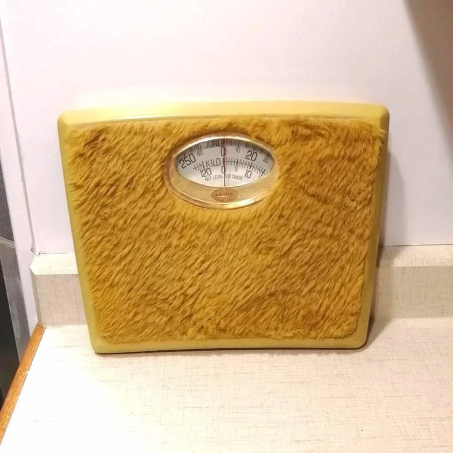 A yellow analog bathroom scale with a fuzzy, textured surface resembling fur, placed on a countertop against a white wall.
