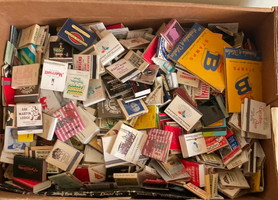 A cardboard box filled with a large, assorted collection of colorful matchbooks, many with different logos, brands, and designs visible, some slightly worn and overlapping each other.