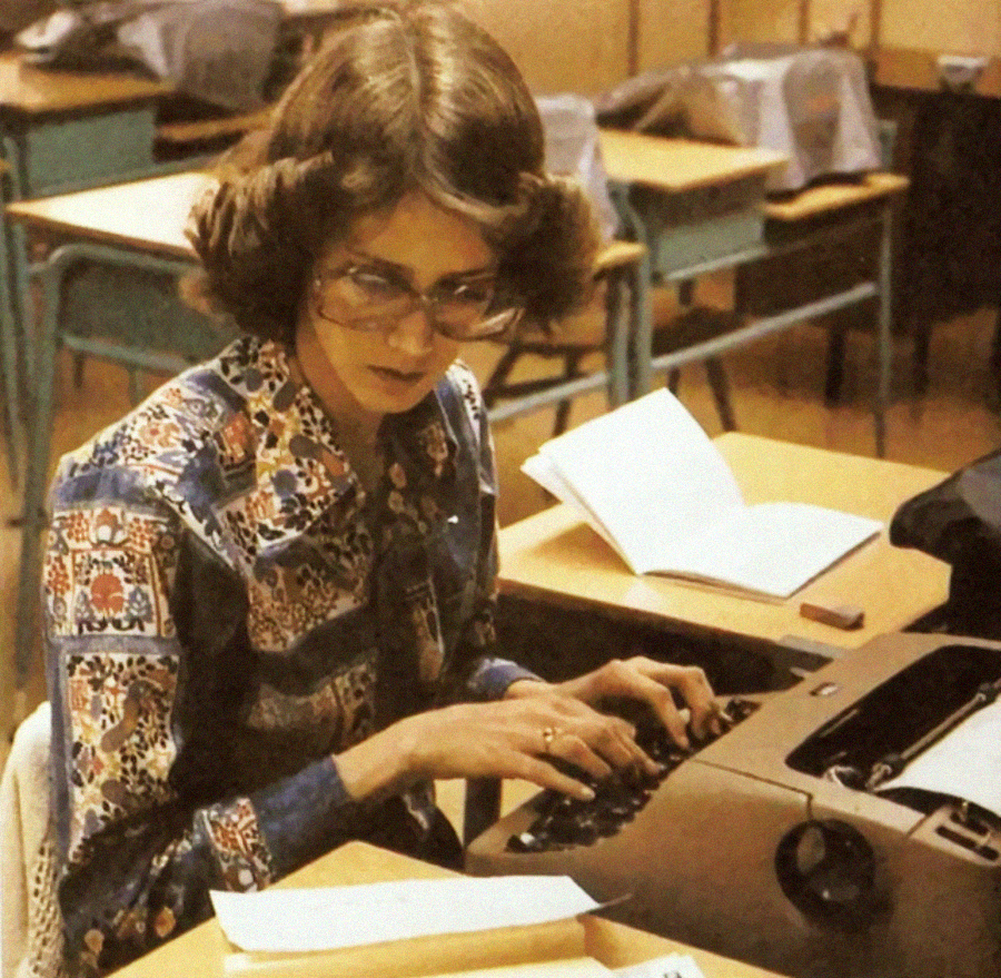 A woman with glasses and a patterned shirt types on a typewriter at a desk in a classroom. Open books and empty chairs are visible in the background.