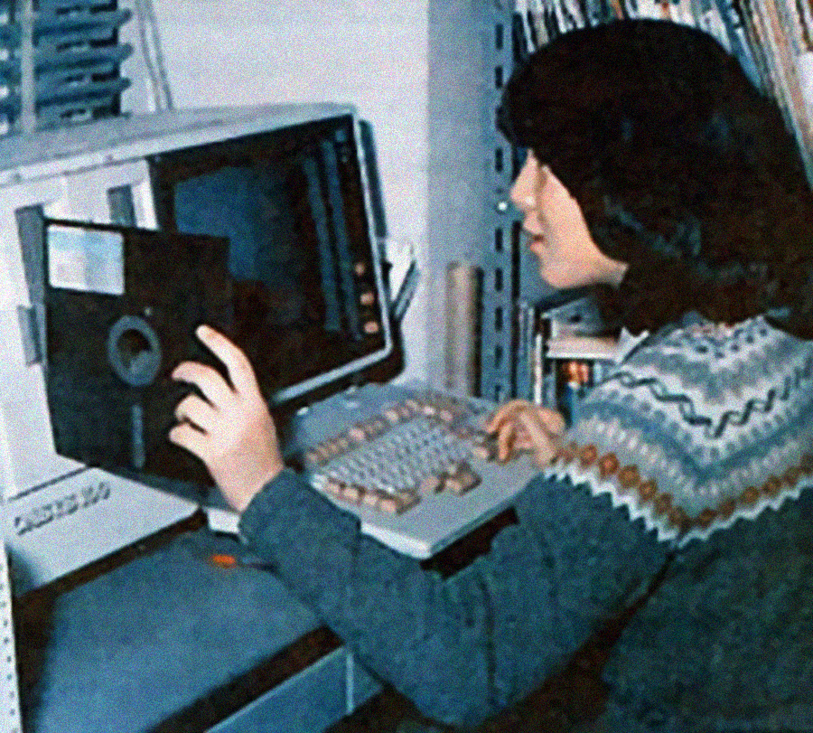 A person with dark hair sits at a vintage computer, inserting a large floppy disk into the disk drive. The individual is wearing a patterned sweater, and shelves with books or files are visible in the background.