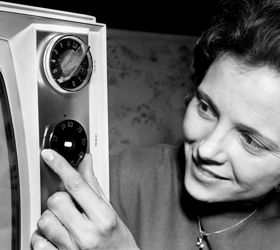 A woman smiles while adjusting the dials on an old-fashioned television set, shown in close-up black-and-white.