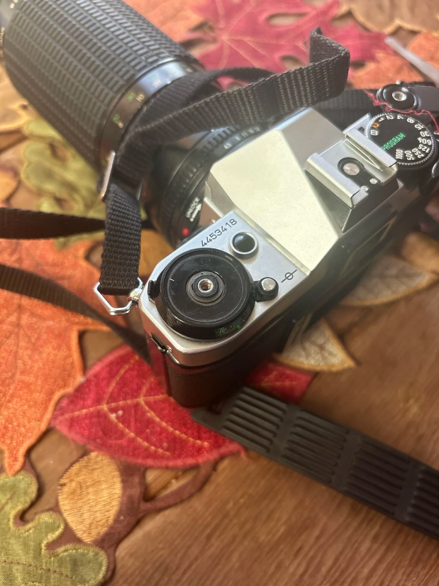 A close-up of a vintage film camera with a long zoom lens and black strap, resting on a colorful autumn-themed tablecloth with leaf patterns.