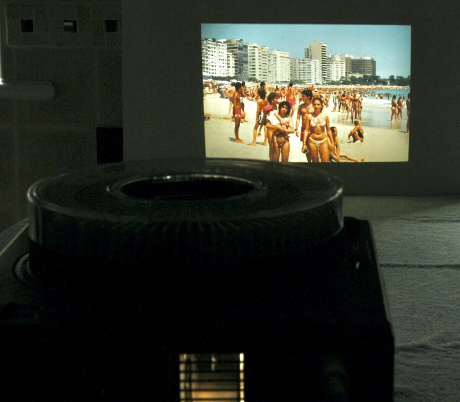 A slide projector displays an image of a crowded beach with many people in swimsuits and tall buildings in the background, projected onto a wall in a dimly lit room.