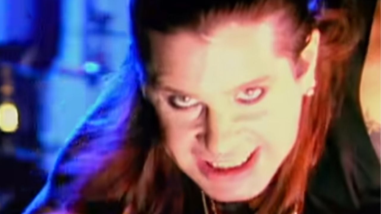 A person with long, reddish-brown hair and intense eye makeup stares forward with a dramatic expression in blue lighting.