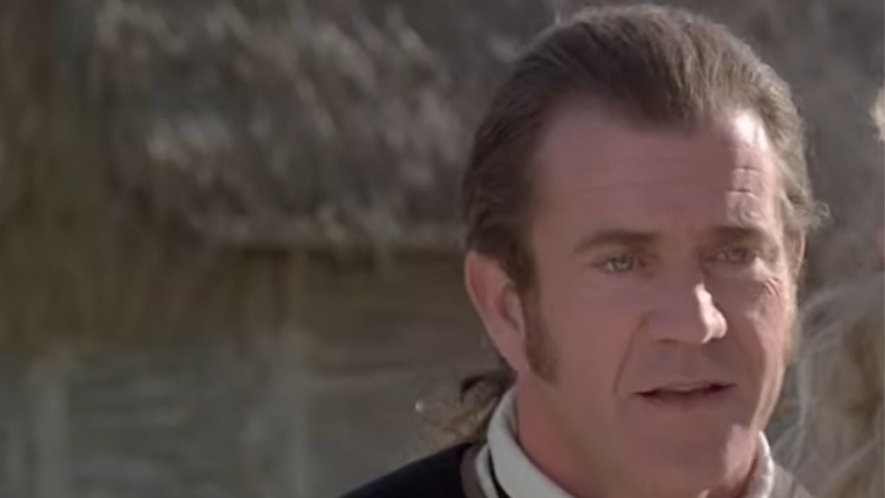 A man with dark hair, sideburns, and period clothing stands outside with a thatched roof building in the background.