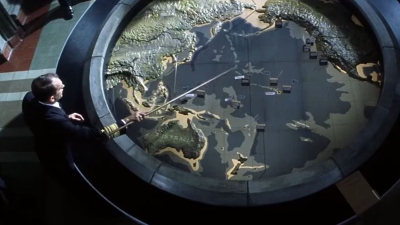 A man in uniform points with a stick at a large, round map table displaying the Pacific region, including parts of Asia, Australia, and the U.S., with markers indicating various locations.