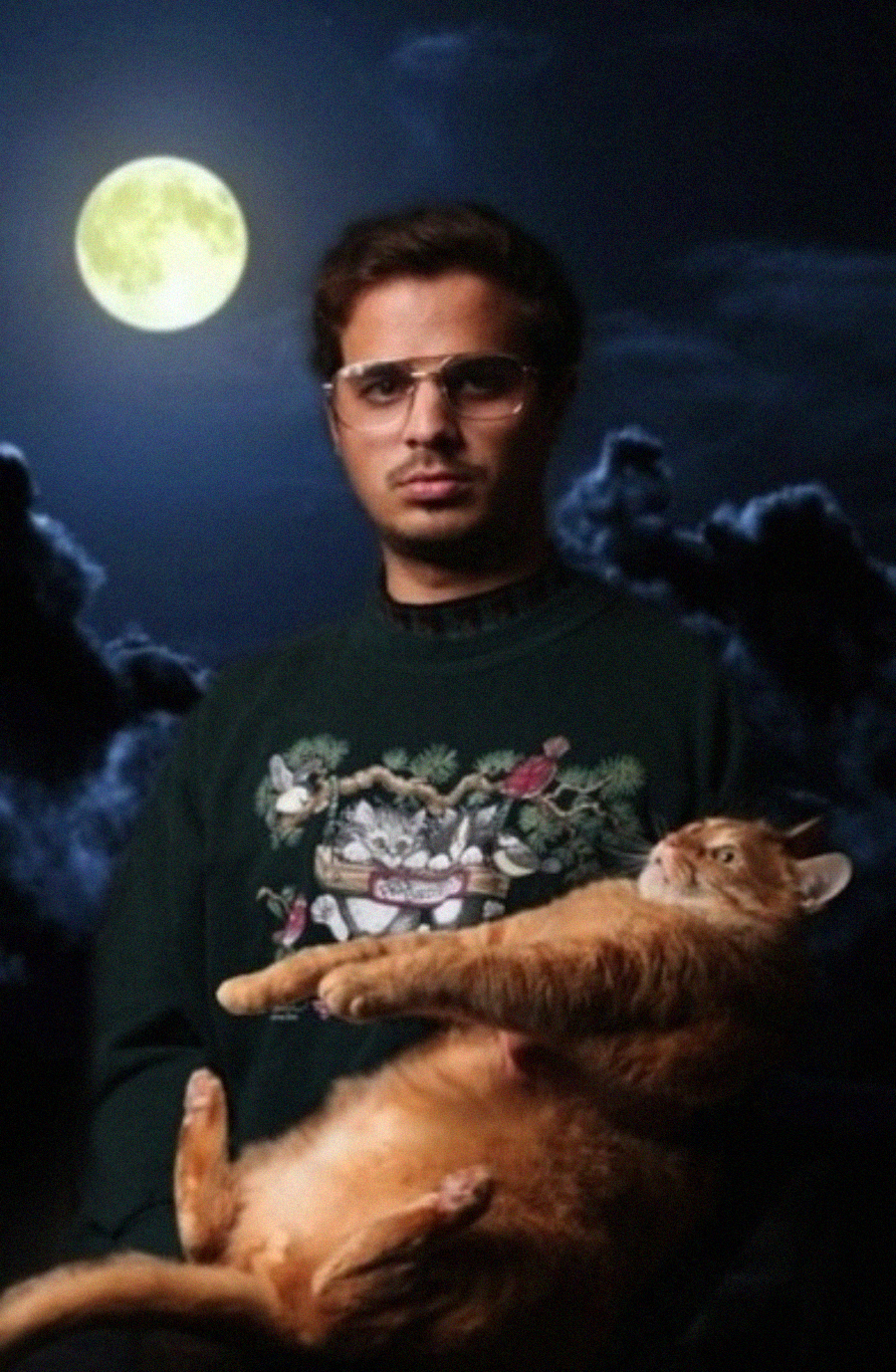 A man wearing glasses and a sweater with a cat design holds a large orange cat in his arms. They are posed in front of a dark, cloudy night sky with a bright full moon.
