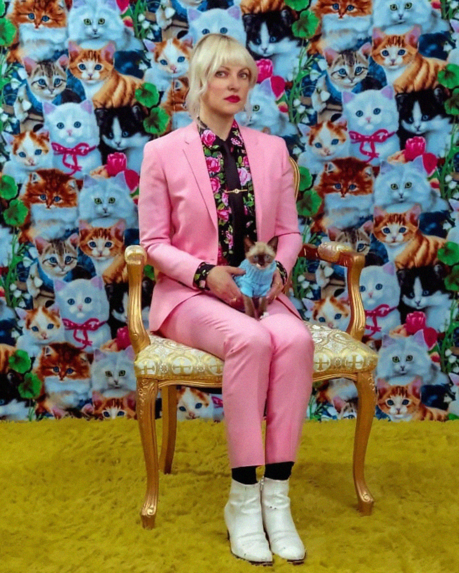 A person in a pink suit and white boots sits on an ornate chair holding a small cat. The background is a colorful, patterned wallpaper covered with various cat faces. The floor is covered with yellow carpet.