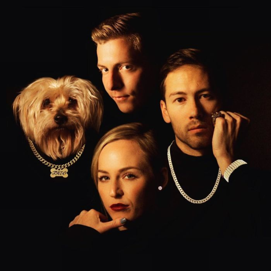 Three people and a dog pose together against a dark background, all wearing black clothes and flashy jewelry. The dog wears a chain necklace with a name tag, and the group looks directly at the camera with serious expressions.