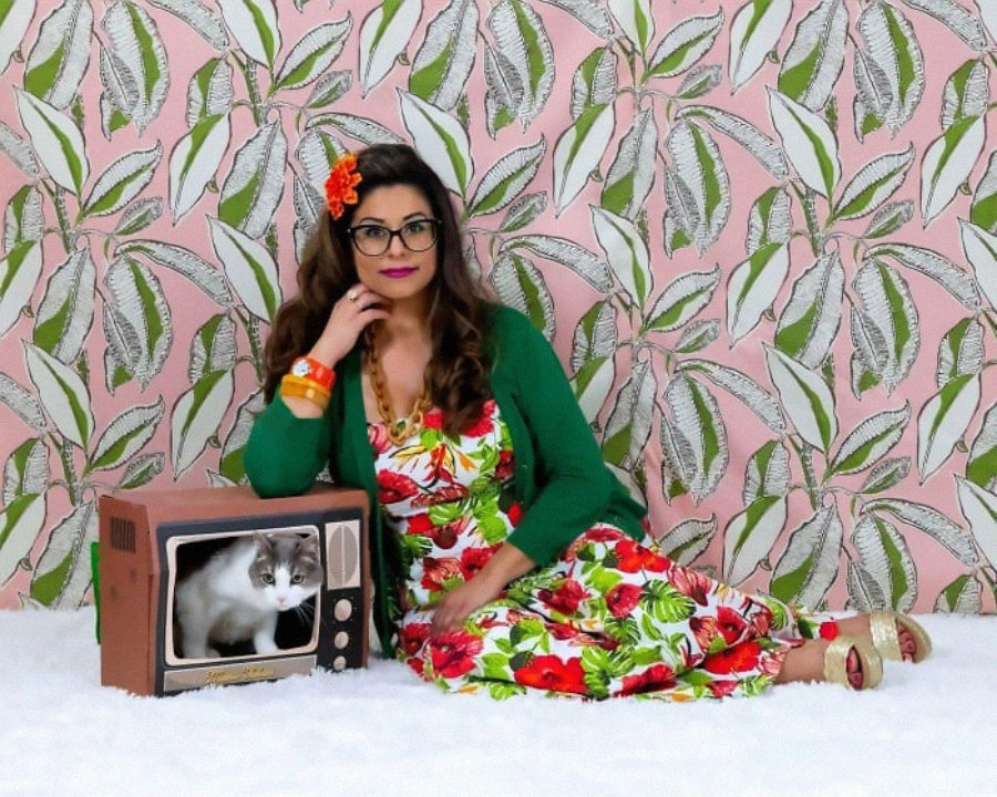 A woman in a colorful floral dress and green cardigan sits on the floor beside a vintage TV with a cat inside. The background is a pink wallpaper with a large leafy pattern.