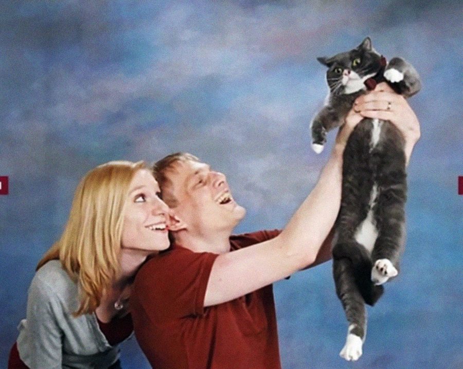 A smiling couple poses for a quirky photo in front of a blue-gray backdrop, with the man holding up a large, grumpy-looking gray and white cat, while the woman looks up at the cat and smiles.