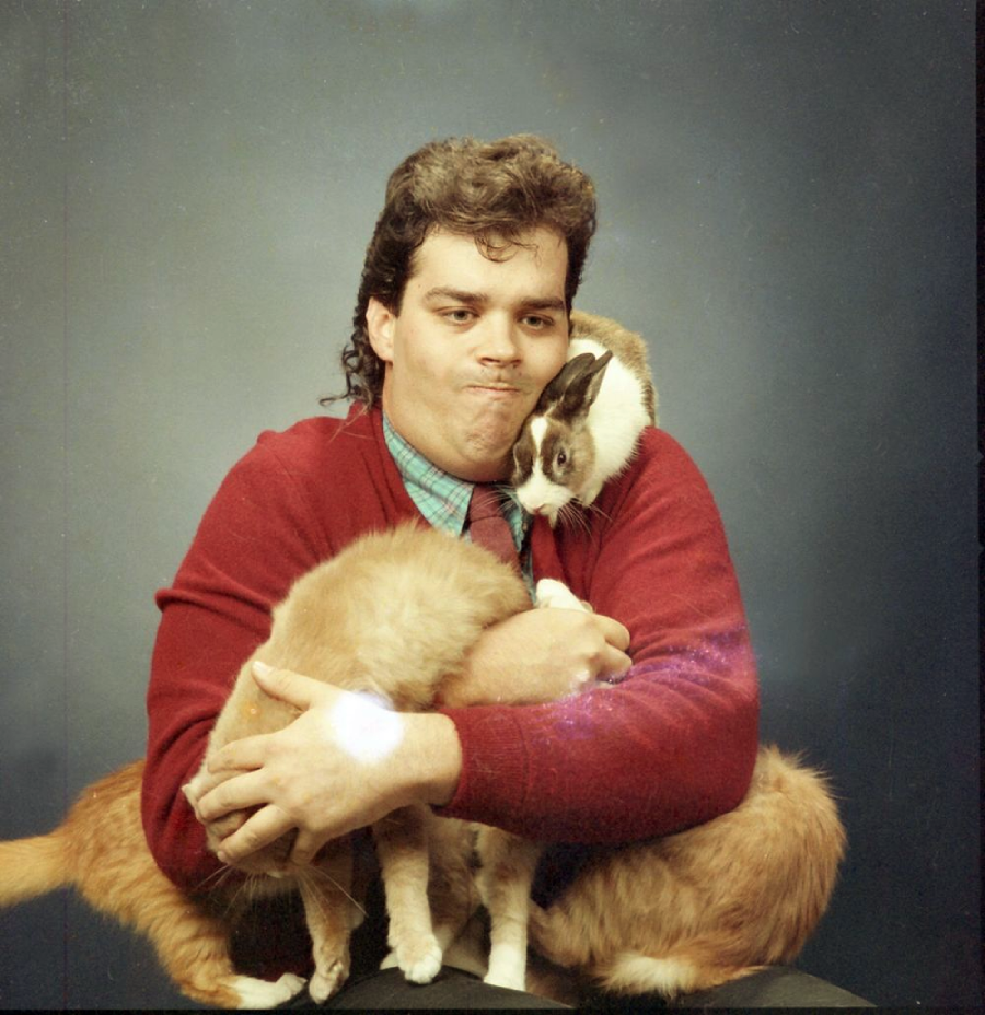 A man in a red sweater poses with a mullet hairstyle, awkwardly holding an orange cat and a brown and white rabbit against a dark studio backdrop.
