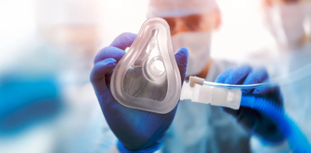 Close-up view of a medical professional in protective gear and blue gloves holding an oxygen mask towards the camera, with blurred background suggesting a clinical or hospital setting.