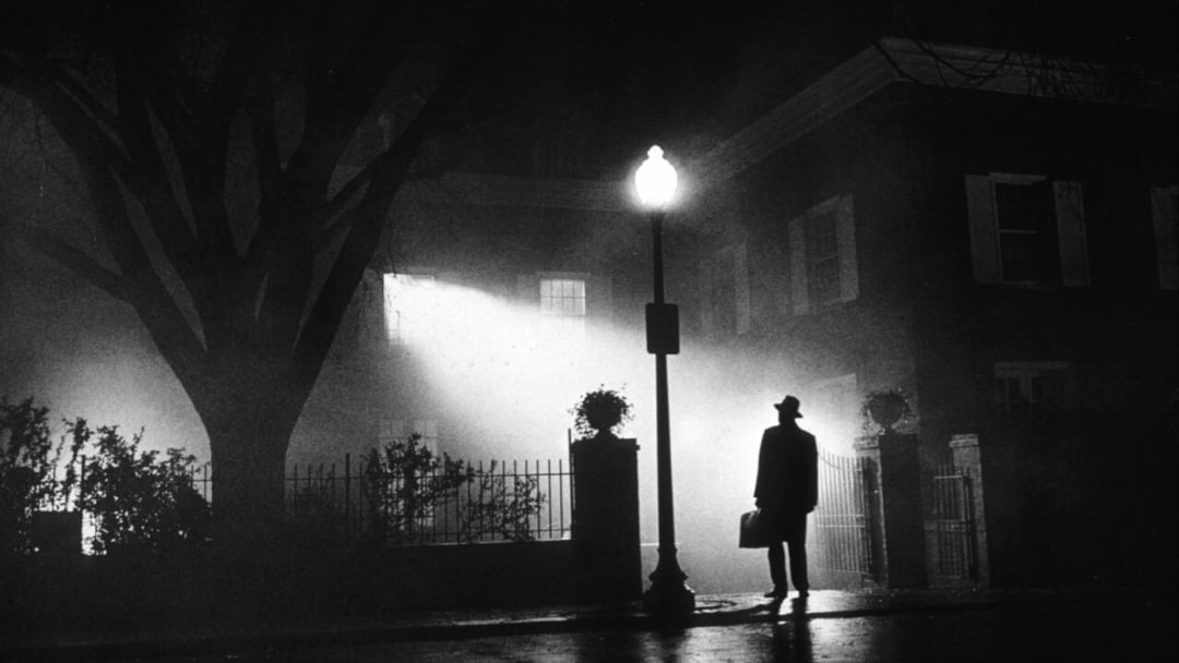 A silhouetted man stands under a streetlamp at night, facing a house with bright light streaming from an upstairs window, casting shadows and creating a mysterious, eerie atmosphere.