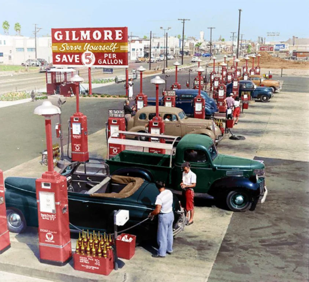 A vintage gas station with red pumps and classic cars. A woman and attendant fill a convertible’s tank. A large sign reads: “Gilmore Serve Yourself, Save 5¢ Per Gallon.” The scene appears from the mid-20th century.