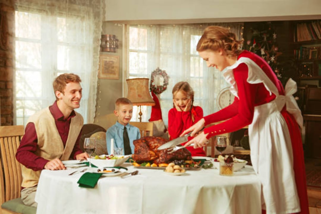 A woman in a red dress and white apron carves a roasted turkey at a festive dining table as a man and two children smile, ready to enjoy the holiday meal in a warmly decorated dining room.