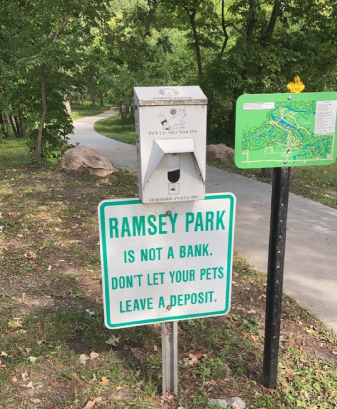A sign at Ramsey Park reads, "Ramsey Park is not a bank. Don't let your pets leave a deposit." Nearby are a bag dispenser for pet waste and a green park map; a paved path winds through trees in the background.