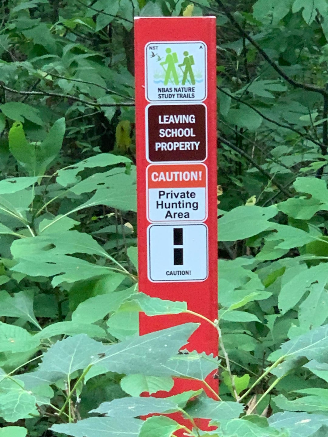 A red trail marker in dense green foliage displays signs reading: "Leaving School Property," "Caution! Private Hunting Area," and "NBAS Nature Study Trails" with caution symbols.