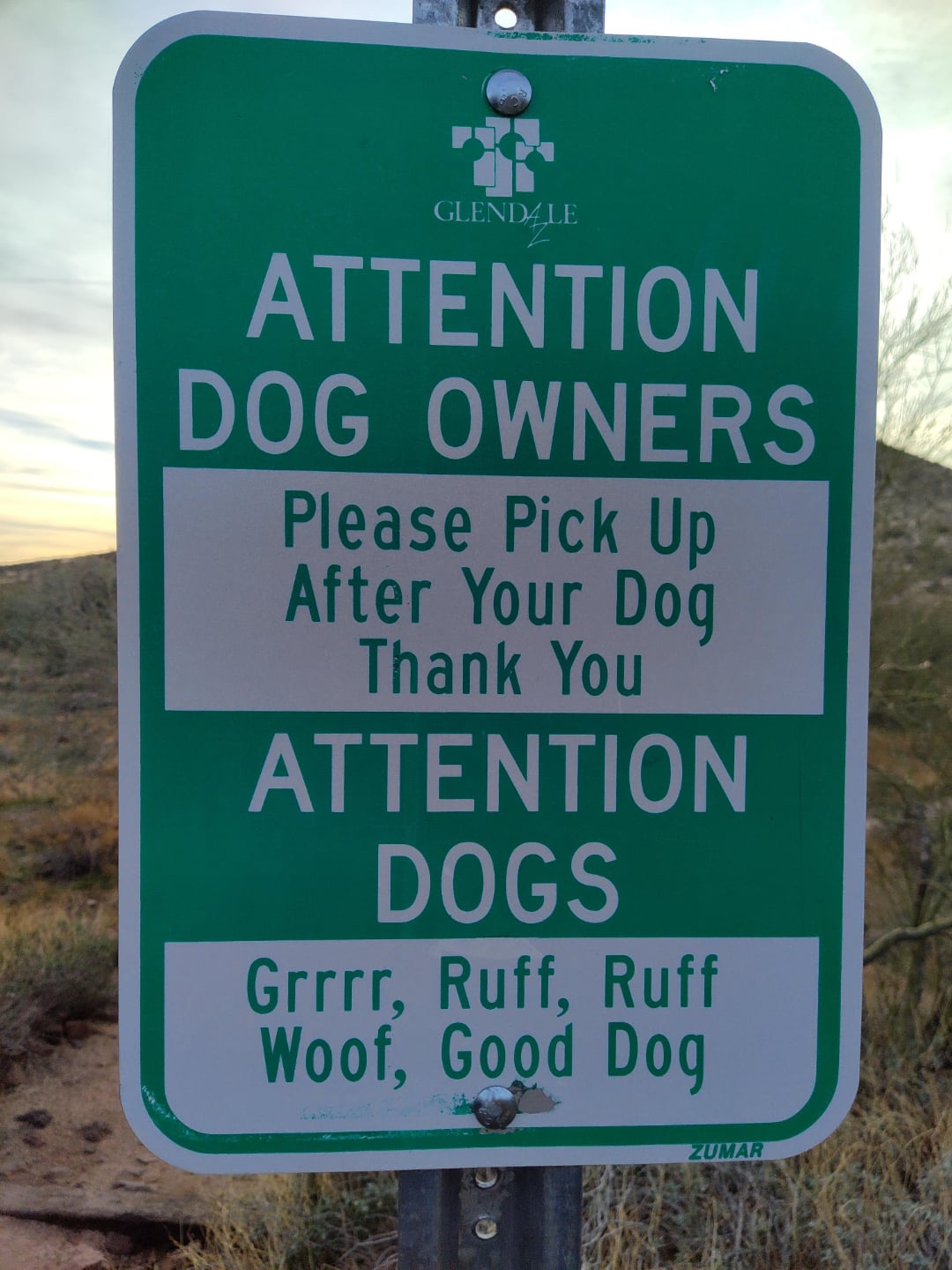 A green and white Glendale park sign reads: “Attention Dog Owners: Please pick up after your dog. Thank you. Attention Dogs: Grrr, Ruff, Ruff, Woof, Good Dog.” Desert landscape in background.