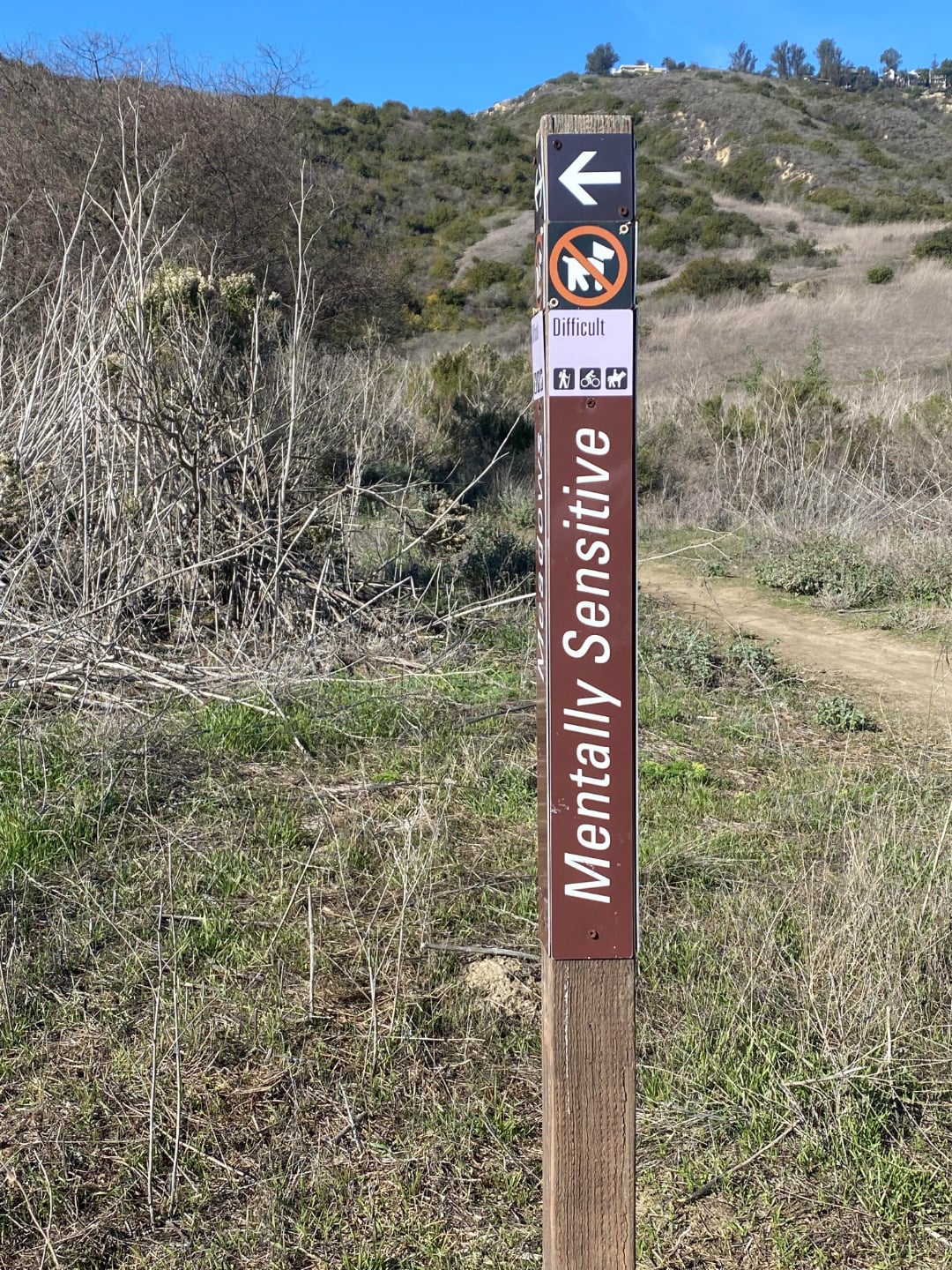 A trail sign in a grassy, hilly area reads "Mentally Sensitive" and points left. It indicates the trail is difficult, no bikes allowed, and features hiking symbols under a clear blue sky.