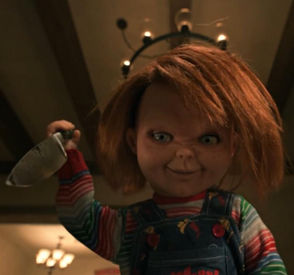 A doll with red hair, wearing striped clothing and denim overalls, holds a large knife and grins menacingly under a ceiling light, creating a sinister atmosphere.