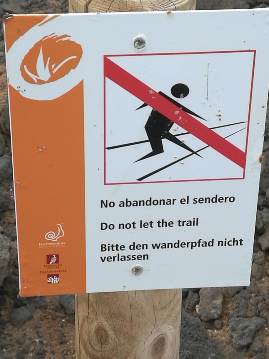 A sign on a wooden post shows a figure walking off a trail with a red line crossing it out. It reads: "No abandonar el sendero. Do not let the trail. Bitte den wanderpfad nicht verlassen.
