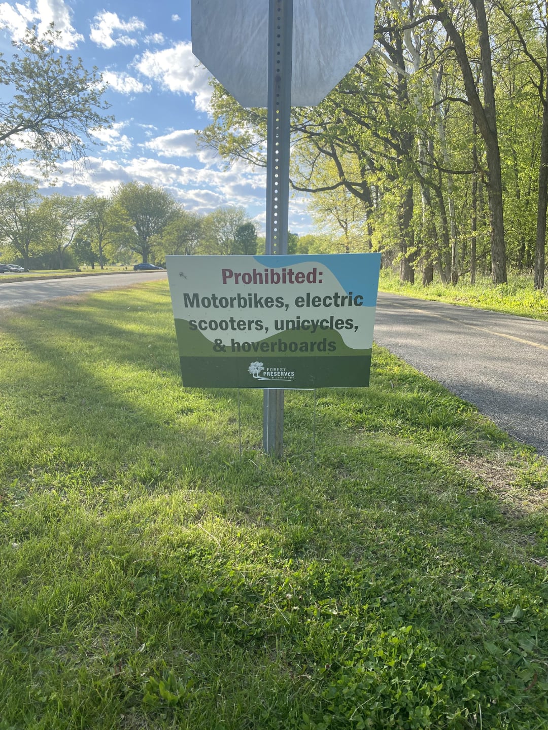 A sign on grass beside a road reads: "Prohibited: Motorbikes, electric scooters, unicycles, & hoverboards." Trees and a partially cloudy sky are visible in the background.