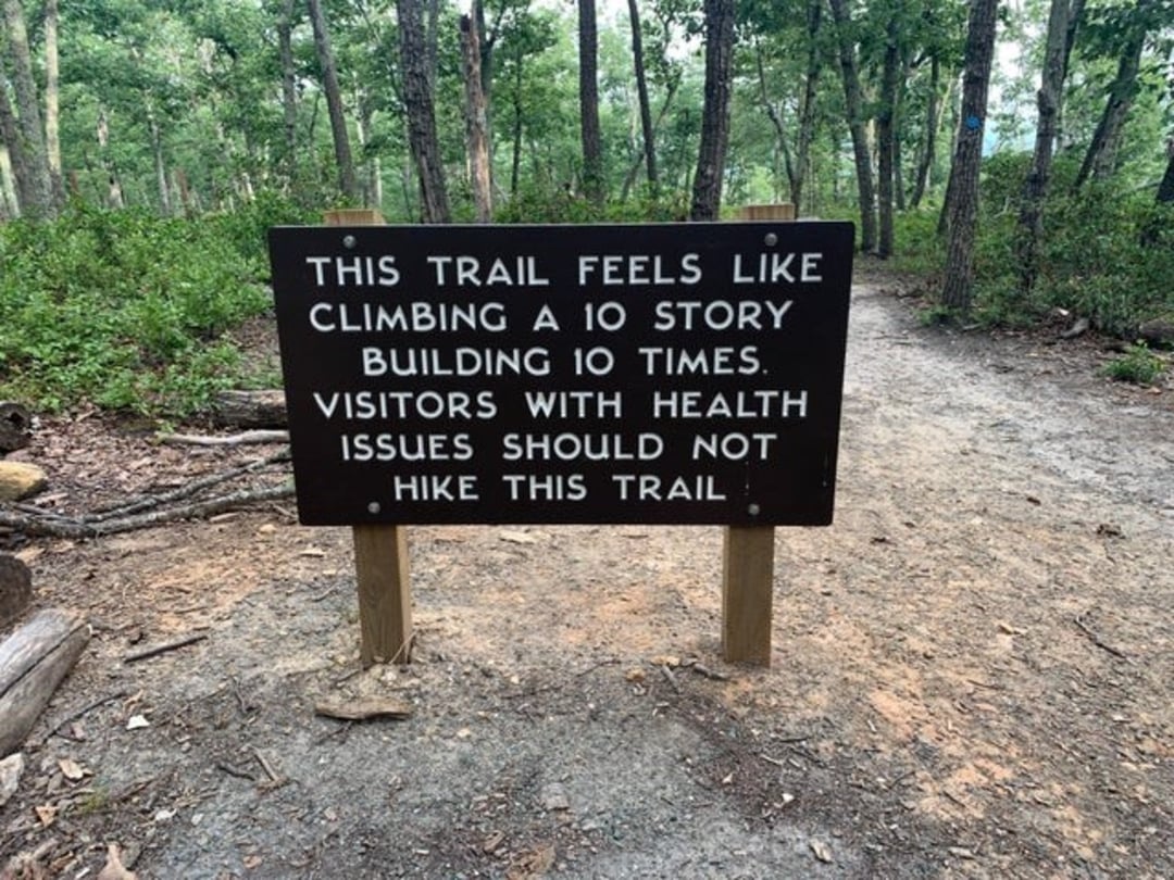 A sign on a forest trail reads: "This trail feels like climbing a 10 story building 10 times. Visitors with health issues should not hike this trail." It stands on a dirt path surrounded by trees.