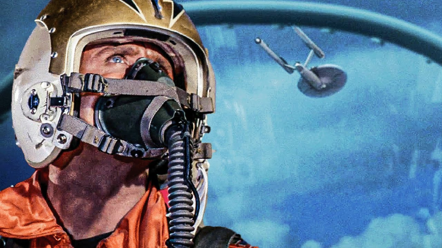 A pilot wearing a gold helmet and oxygen mask looks up from a cockpit, with the Starship Enterprise visible outside against a blue sky.