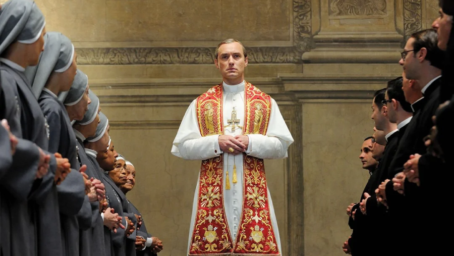 A man dressed as a pope stands solemnly in the center of an ornate hall, flanked by nuns on one side and priests on the other, all looking toward him with serious expressions.