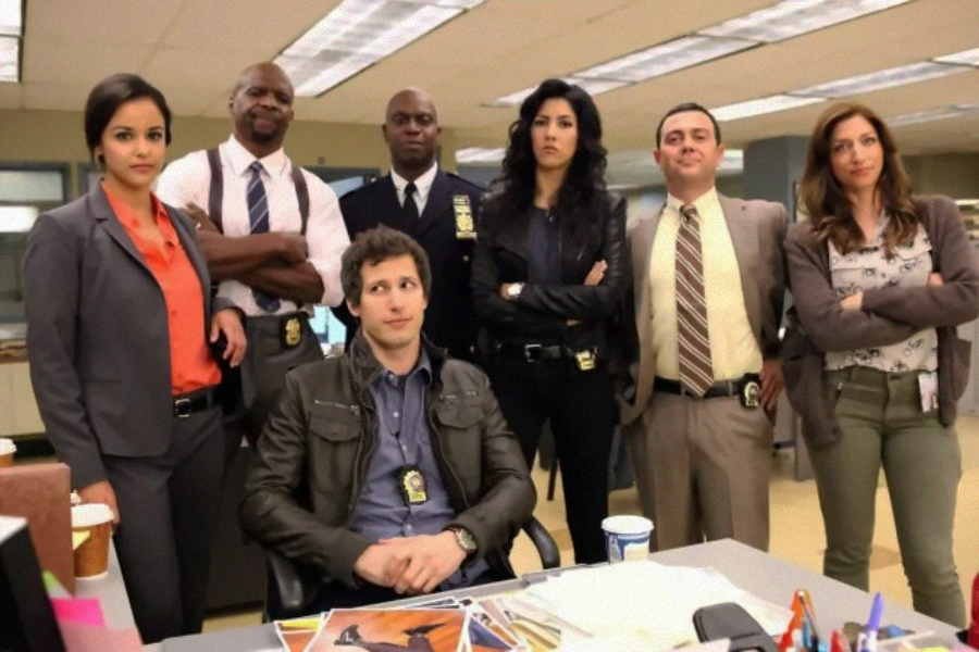 Seven people, dressed in business attire and police badges, stand and sit in an office setting. Six stand with arms crossed behind a seated man at a cluttered desk. They appear confident and professional.